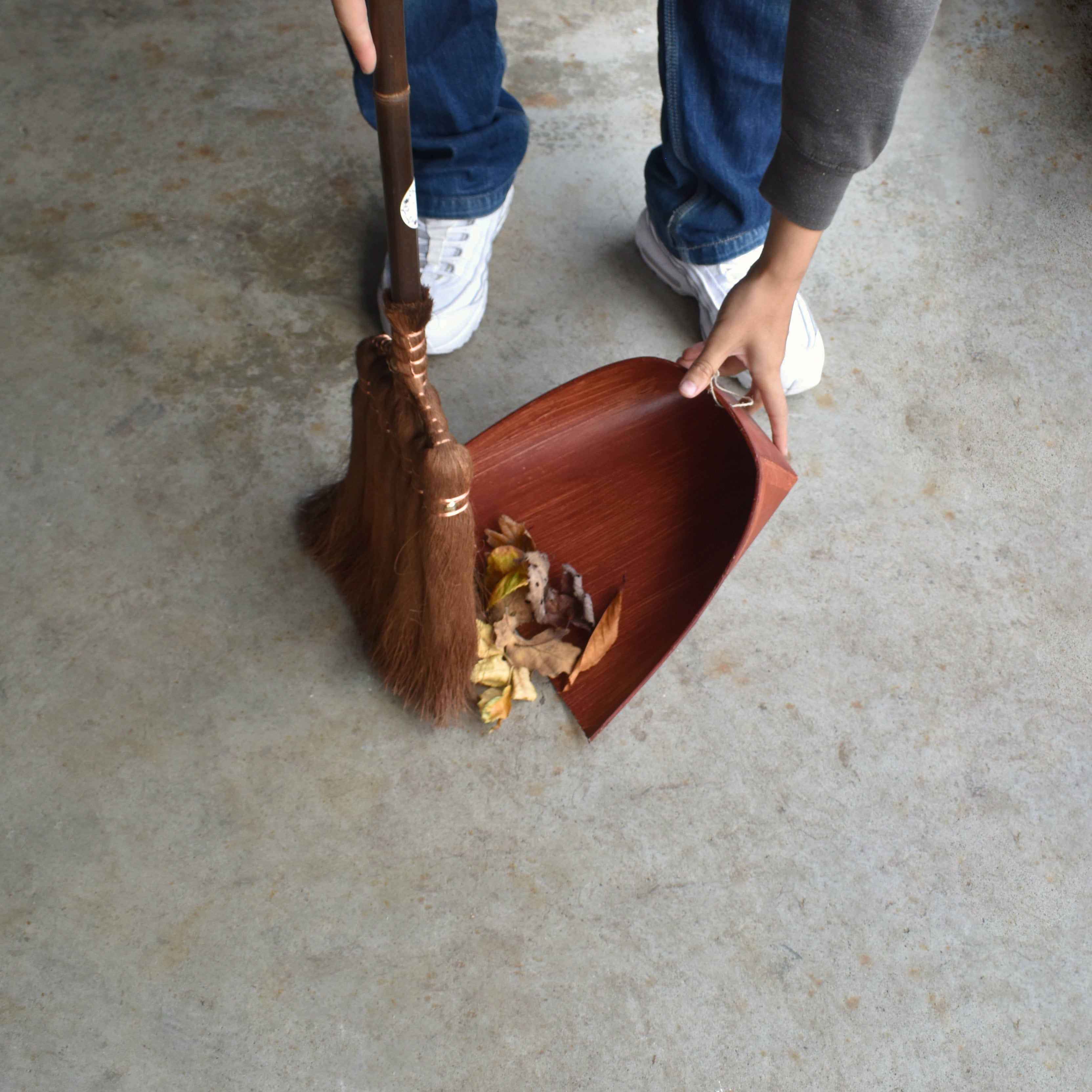 Person sweeping fallen leaves into a traditional Japanese shuro broom and handcrafted wooden dustpan on a concrete floor.
