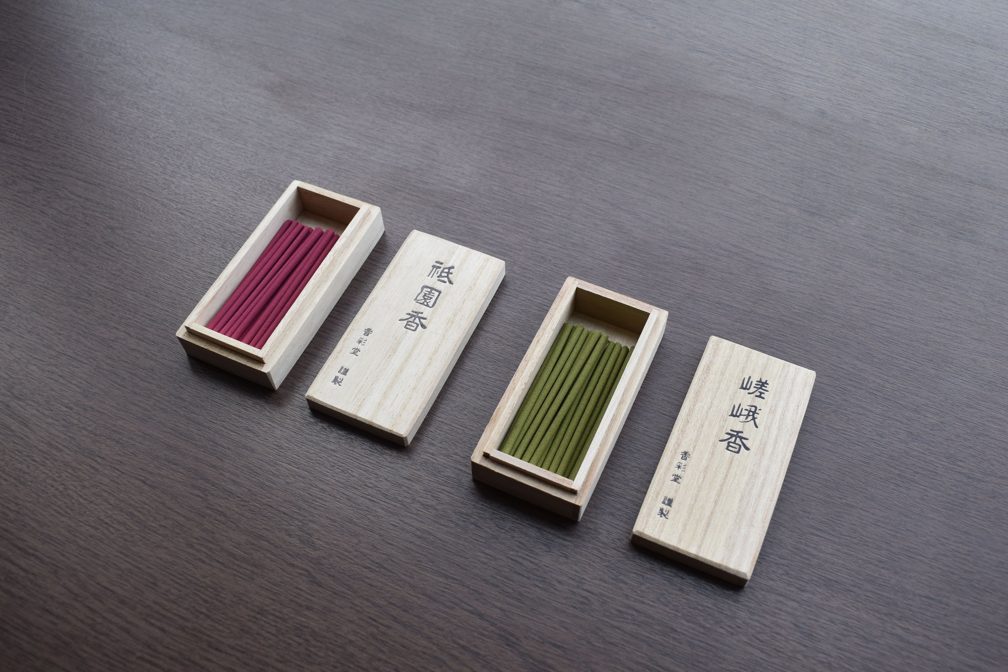Two open wooden boxes of Japanese incense sticks—one red and one green—are displayed neatly on a dark wooden surface, with their respective engraved lids placed beside each box.