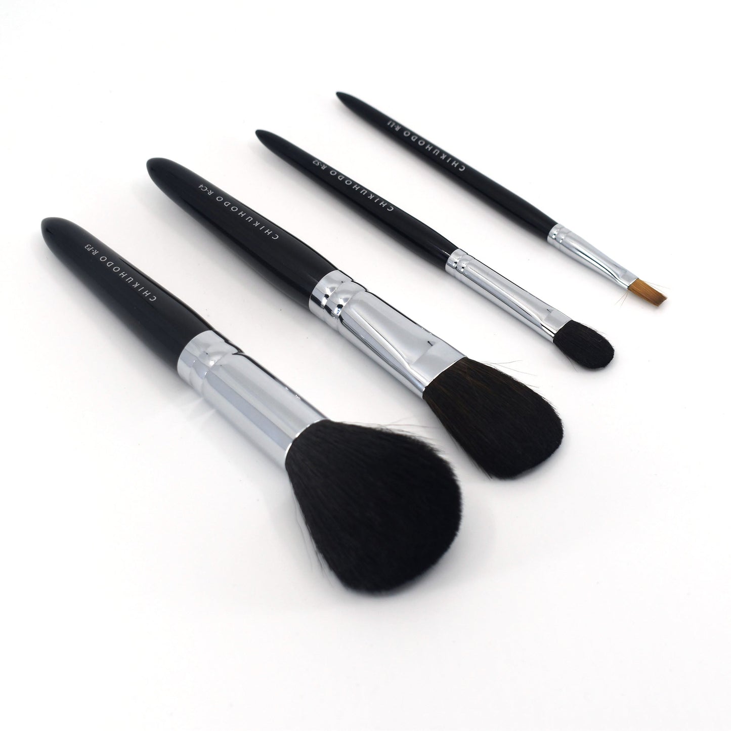 Angled view of four Chikuhodo makeup brushes arranged diagonally on a white surface, featuring sleek black handles, silver ferrules, and soft bristles in varying shapes for face and eye application.