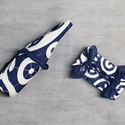 Two furoshiki-wrapped gifts displayed on a grey surface — one bottle and one rectangular box wrapped in indigo blue and white traditional Japanese cloth with modern swirl patterns.