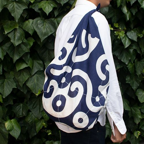 person wearing a traditional Japanese furoshiki cloth bag with bold indigo and white swirl pattern, tied securely over one shoulder, standing against a backdrop of lush green ivy