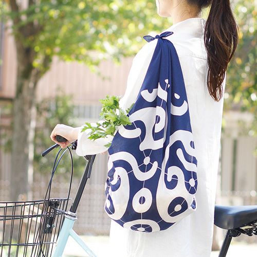 Japanese woman riding a bicycle with a reusable indigo furoshiki bag tied over her shoulder, featuring a bold white swirl pattern and fresh herbs peeking out.