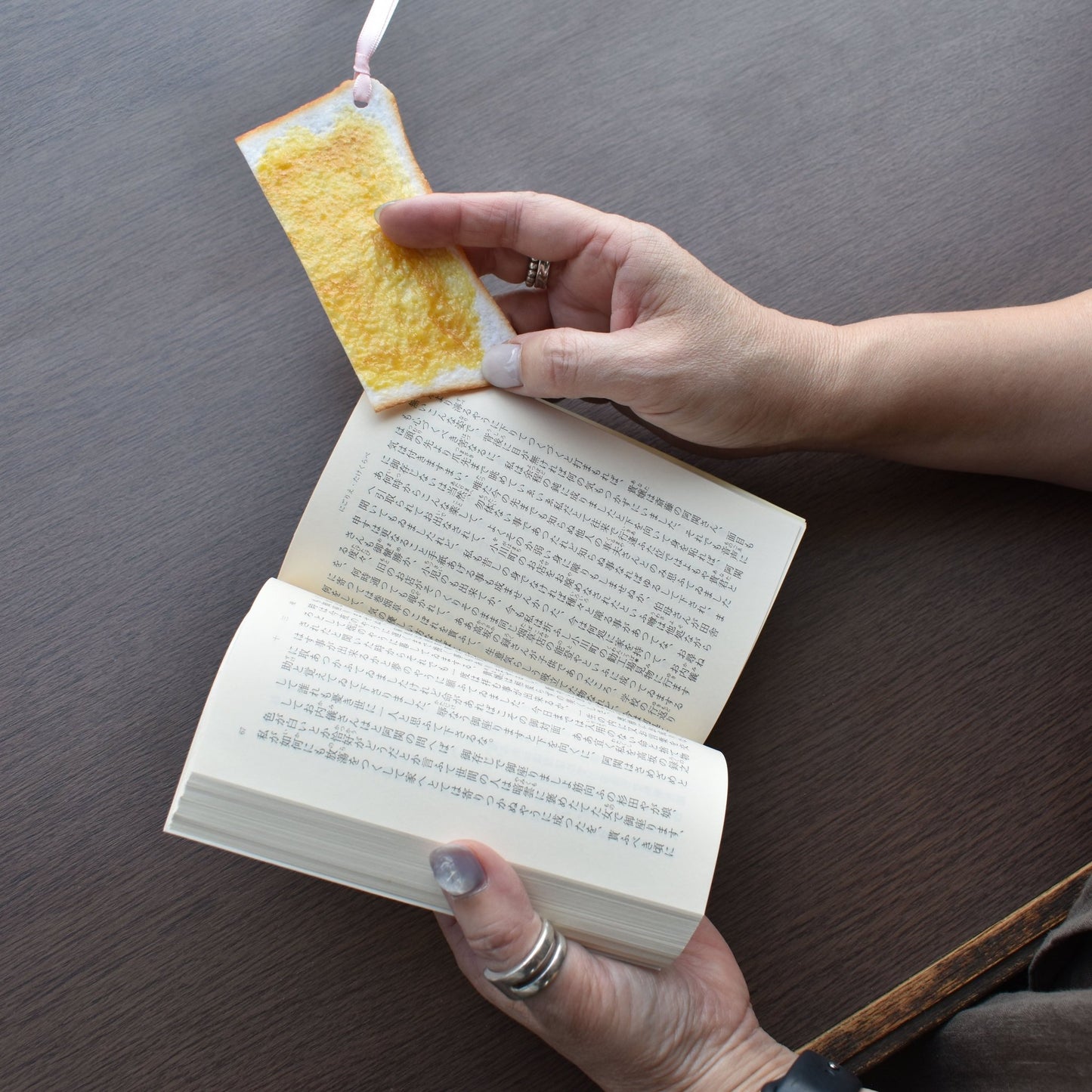 Toast Bookmark. A person holding a book and using a hyper-realistic toast-shaped bookmark made by Nagao Shokken, a Japanese artisan known for food sample-inspired accessories.”