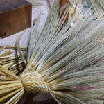 Partially assembled Edo-style broom showing the bound and braided base with splayed broomcorn stalks before trimming. The image captures the intricate weave pattern and the raw, uncut fibers during the early stage of traditional broom making in Japan. Tools and bamboo stalks are visible in the background, highlighting the workshop setting.