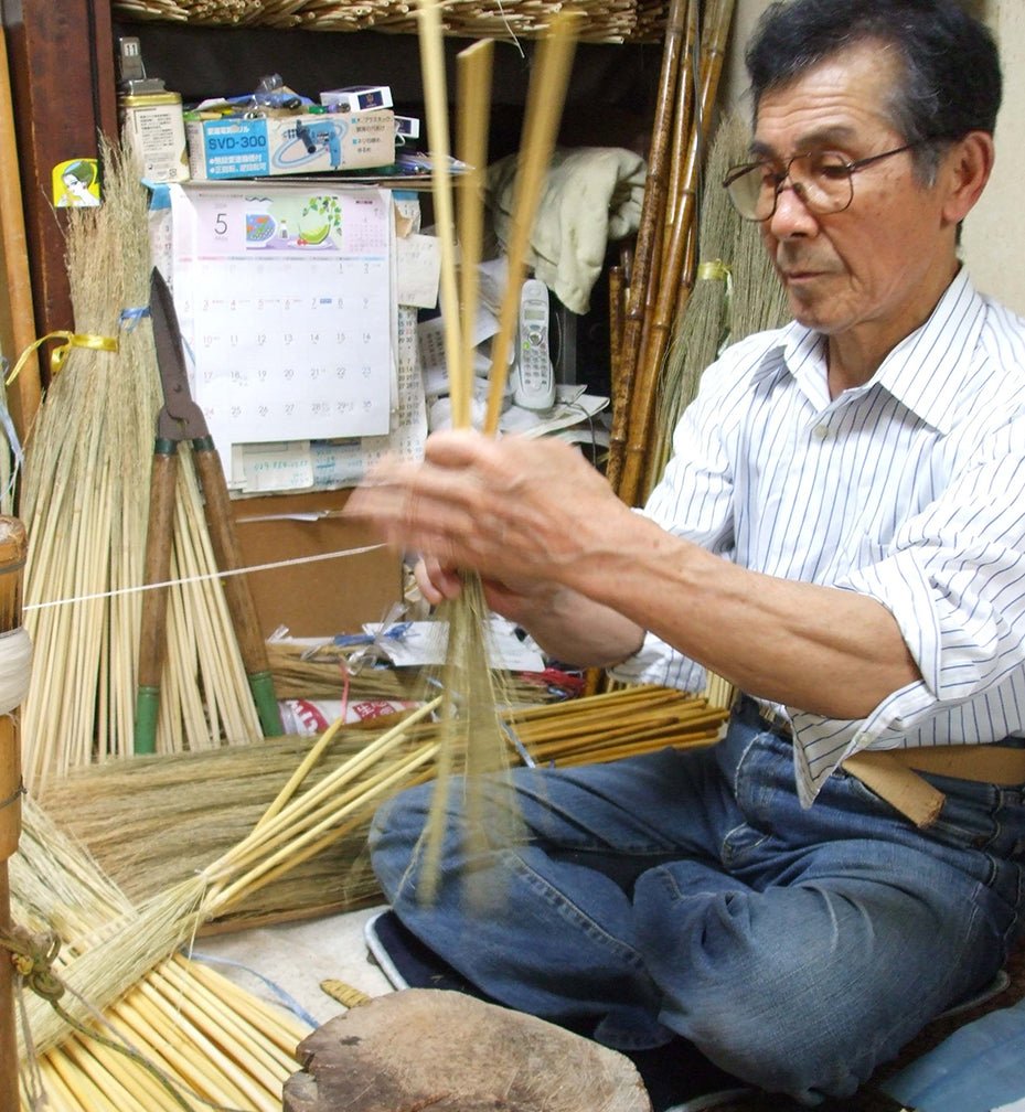 A Japanese artisan skillfully handcrafting an Edo-style broom using traditional materials and techniques. The craftsman sits cross-legged, carefully weaving broomcorn with twine in a cluttered workshop filled with tools, bamboo handles, and handmade brooms in progress—highlighting the meticulous artistry behind Shirokiya Denbei brooms.