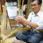 A Japanese artisan skillfully handcrafting an Edo-style broom using traditional materials and techniques. The craftsman sits cross-legged, carefully weaving broomcorn with twine in a cluttered workshop filled with tools, bamboo handles, and handmade brooms in progress—highlighting the meticulous artistry behind Shirokiya Denbei brooms.