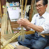 A Japanese artisan skillfully handcrafting an Edo-style broom using traditional materials and techniques. The craftsman sits cross-legged, carefully weaving broomcorn with twine in a cluttered workshop filled with tools, bamboo handles, and handmade brooms in progress—highlighting the meticulous artistry behind Shirokiya Denbei brooms.