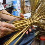 A Japanese craftsman skillfully works on the handle of a traditional Edo-style broom, securing bamboo stalks with string and tightening the structure by hand. The image highlights the intricate braiding and careful knife work involved in assembling the upper section of the broom, surrounded by tools and materials in a cluttered but authentic workshop setting.