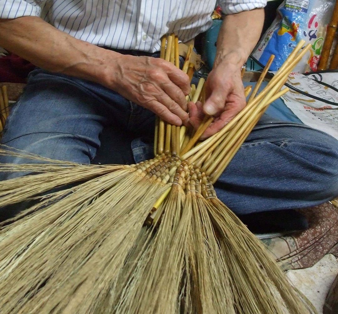 A close-up view of a Japanese artisan weaving the bamboo handle of a traditional Edo-style broom. The craftsman’s hands are carefully threading stalks into the central structure, showcasing the intricate and time-honored technique of handcrafting brooms with precision and care in a compact workshop environment.