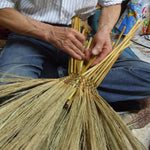 A close-up view of a Japanese artisan weaving the bamboo handle of a traditional Edo-style broom. The craftsman’s hands are carefully threading stalks into the central structure, showcasing the intricate and time-honored technique of handcrafting brooms with precision and care in a compact workshop environment.