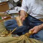 Close-up view of a Japanese craftsman weaving a traditional Edo-style broom by hand. The artisan holds the broom head while shaping and securing the broomcorn stalks with a wooden mallet, surrounded by raw materials and tools in a compact, cluttered workshop. This scene highlights the detailed craftsmanship and tactile process behind handmade Shirokiya Denbei brooms.