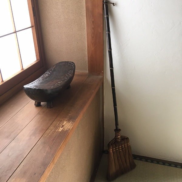 A full-length traditional Japanese shuro broom with a dark bamboo handle resting upright in the corner of a tatami room, beside a wooden windowsill with a carved wooden seat, creating a serene, minimalist interior.