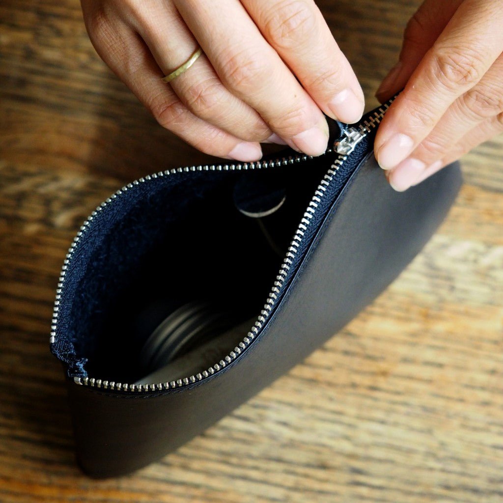 Close-up of hands unzipping a sleek black leather pouch on a wooden surface, revealing storage space for daily essentials.