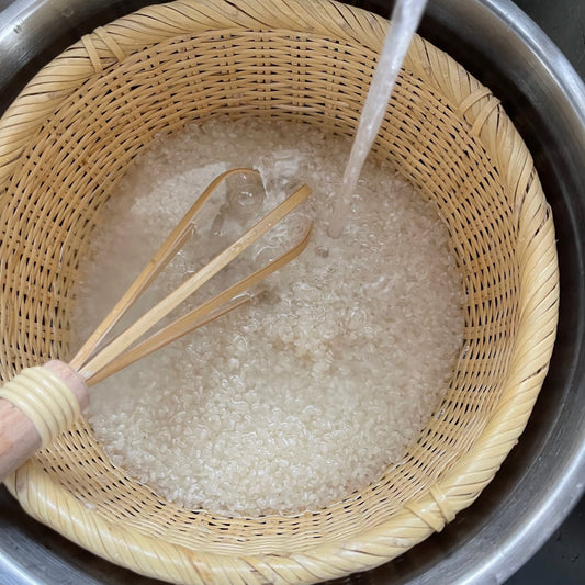 Handwashing rice in a Japanese bamboo basket under running water using a bamboo whisk, showing the basket’s natural drainage and traditional kitchen use
