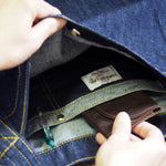 Hand placing wallet into inner snap pocket of Japanese denim shoulder bag, showing organized interior with pen slot and brand label. 