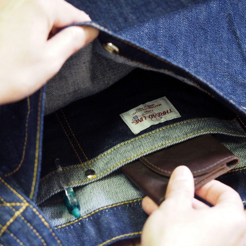 Hand placing wallet into inner snap pocket of Japanese denim shoulder bag, showing organized interior with pen slot and brand label. 