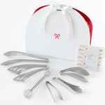 Japanese Precision Kitchen Set, the white half-moon gift box with a red ribbon and the six stainless tools fanned out in front beside the cotton cloth.