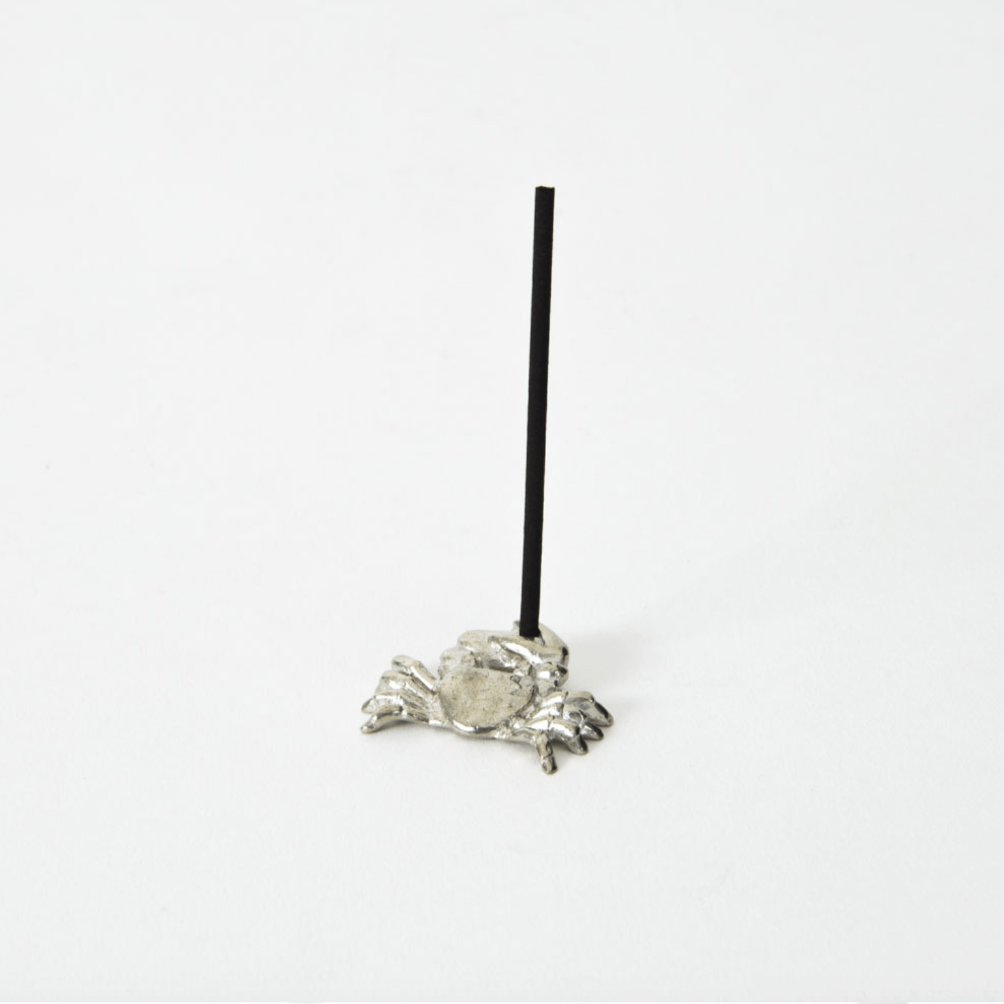 Side view of a silver crab-shaped incense holder with a black incense stick standing upright in the center, placed on a white background.