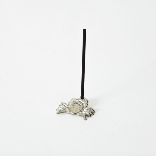 Side view of a silver crab-shaped incense holder with a black incense stick standing upright in the center, placed on a white background.