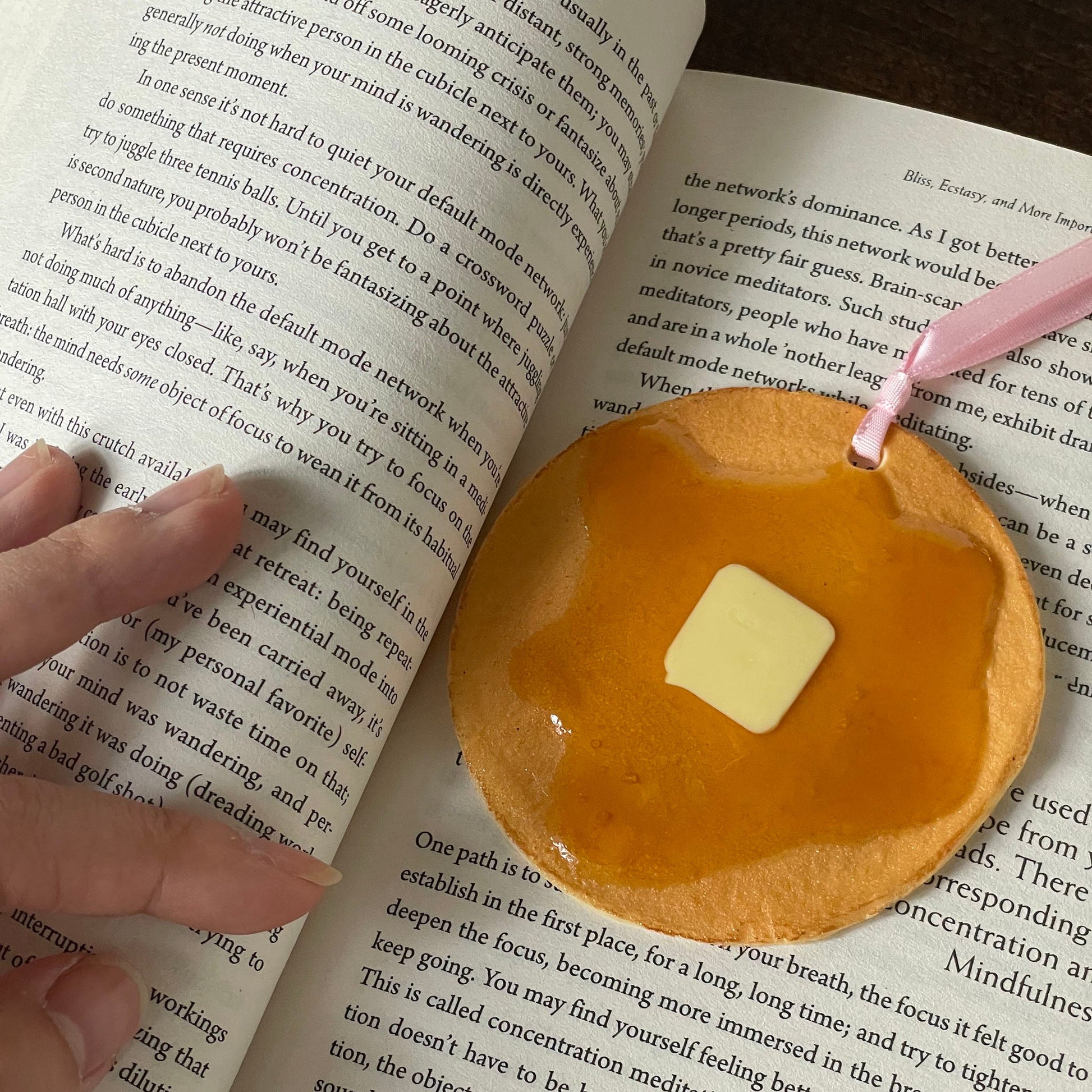 Pancake-shaped resin bookmark with a pat of butter and glossy syrup, resting between the pages of an open book with a pink ribbon attached.