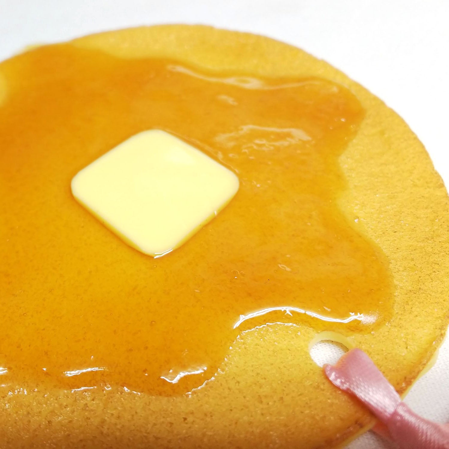 Close-up of a round resin pancake bookmark featuring glossy syrup, a butter pat, and a pink ribbon threaded through the corner.