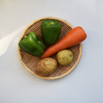 Round bamboo zaru colander holding two green bell peppers, a carrot, and two potatoes on a white surface, showing its functionality for prepping fresh produce.