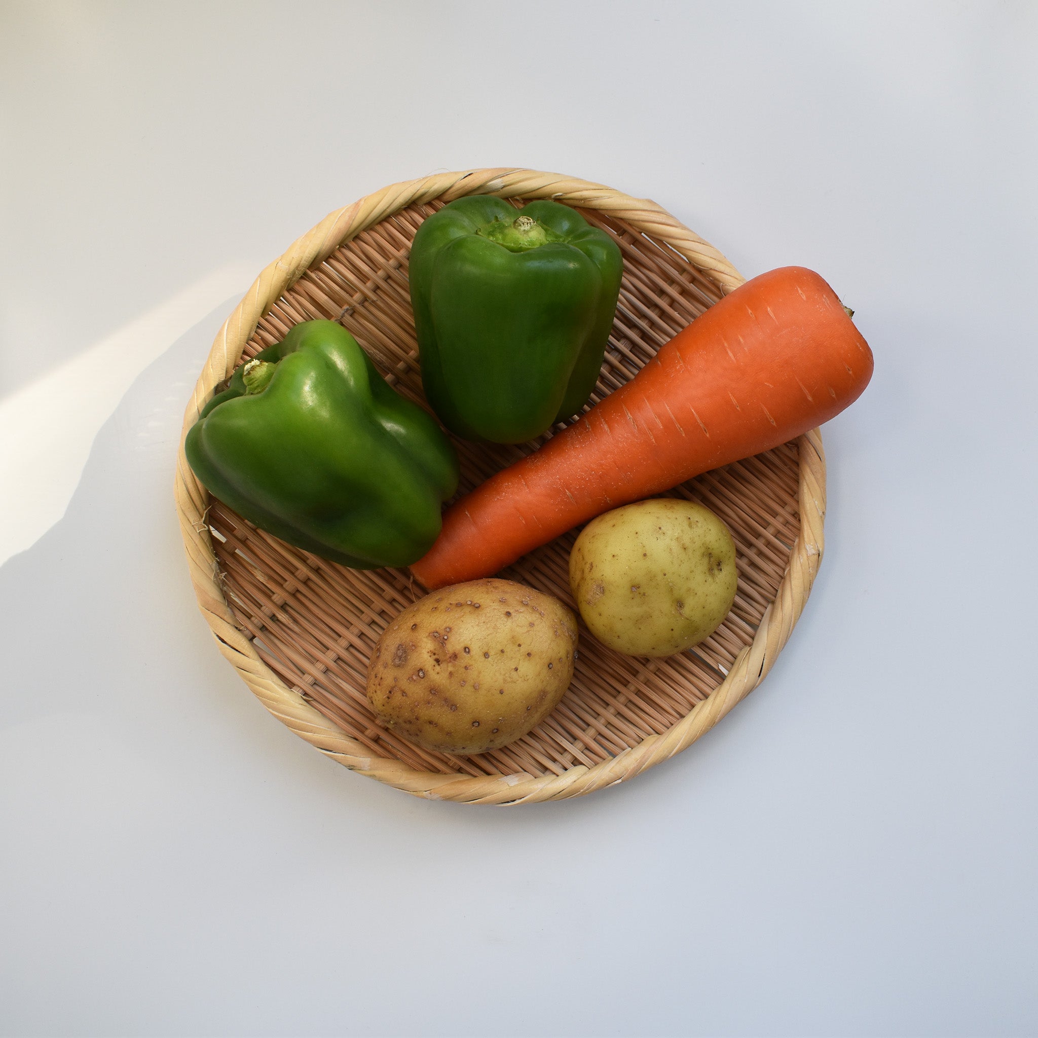 Round bamboo zaru colander holding two green bell peppers, a carrot, and two potatoes on a white surface, showing its functionality for prepping fresh produce.