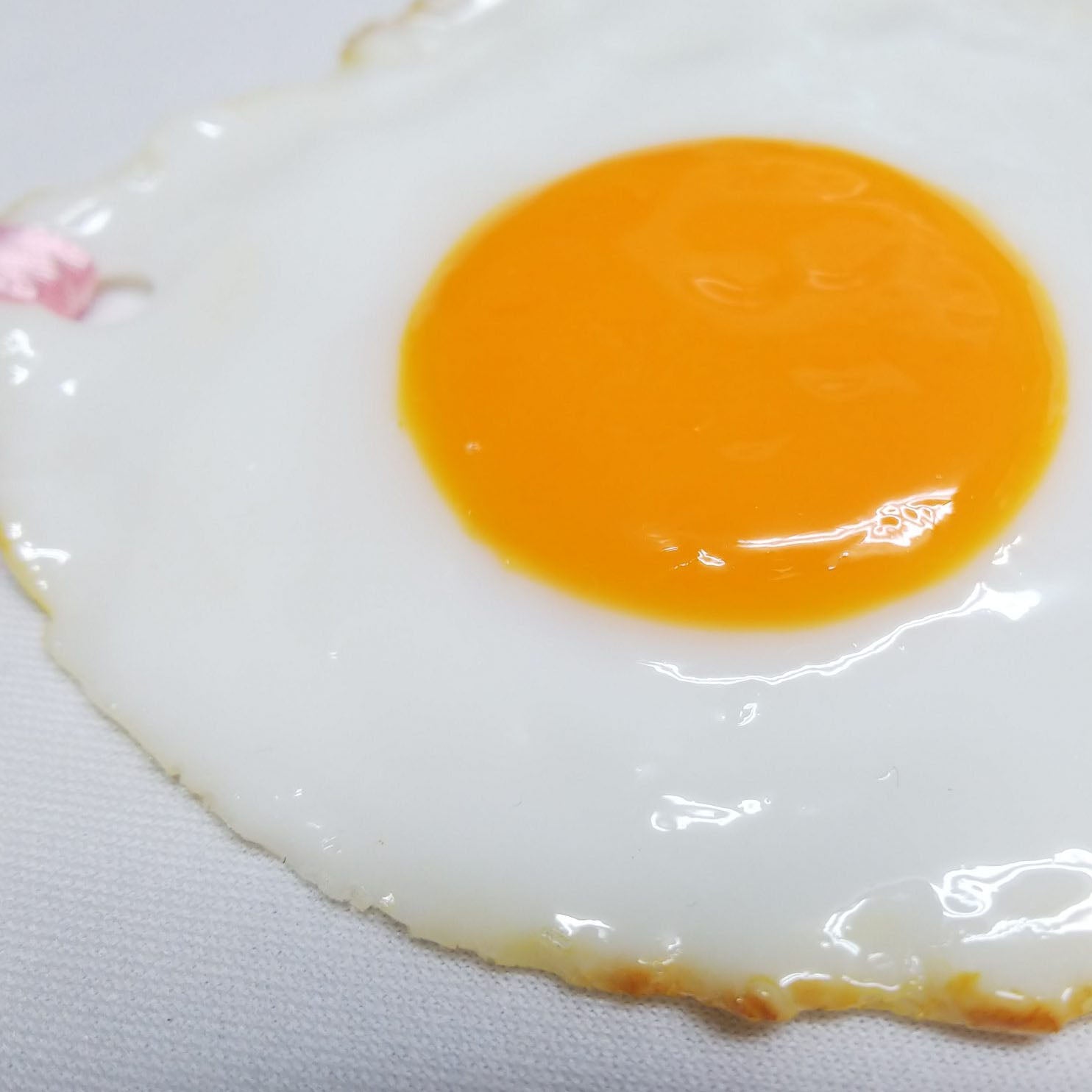 Close-up of a glossy fried egg bookmark showing a vibrant yellow yolk and realistic texture on white fabric background.