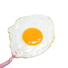 Close-up of a hyper-realistic sunny side up egg bookmark with glossy yolk and white, featuring a pink ribbon loop for easy use.
