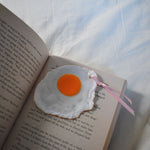 A playful sunny side up egg bookmark resting on an open book, with a glossy yolk and pink ribbon peeking out over soft white bedding.