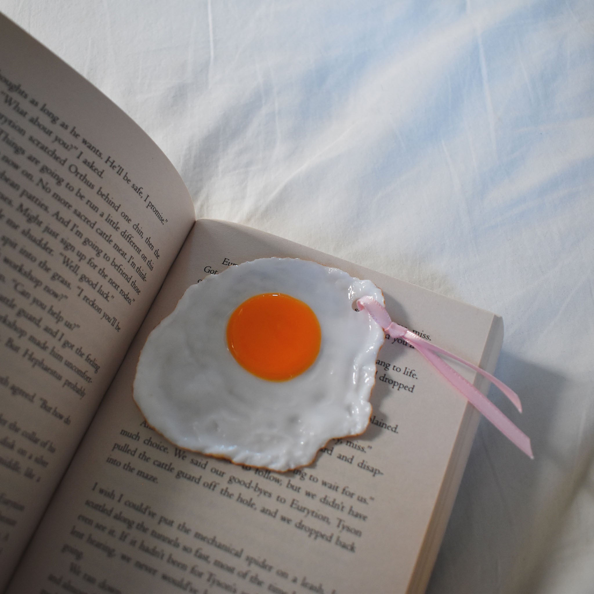 A playful sunny side up egg bookmark resting on an open book, with a glossy yolk and pink ribbon peeking out over soft white bedding.