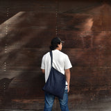 Man wearing a minimalist Japanese denim shoulder bag against a rustic wooden wall, showcasing casual and versatile streetwear style. 