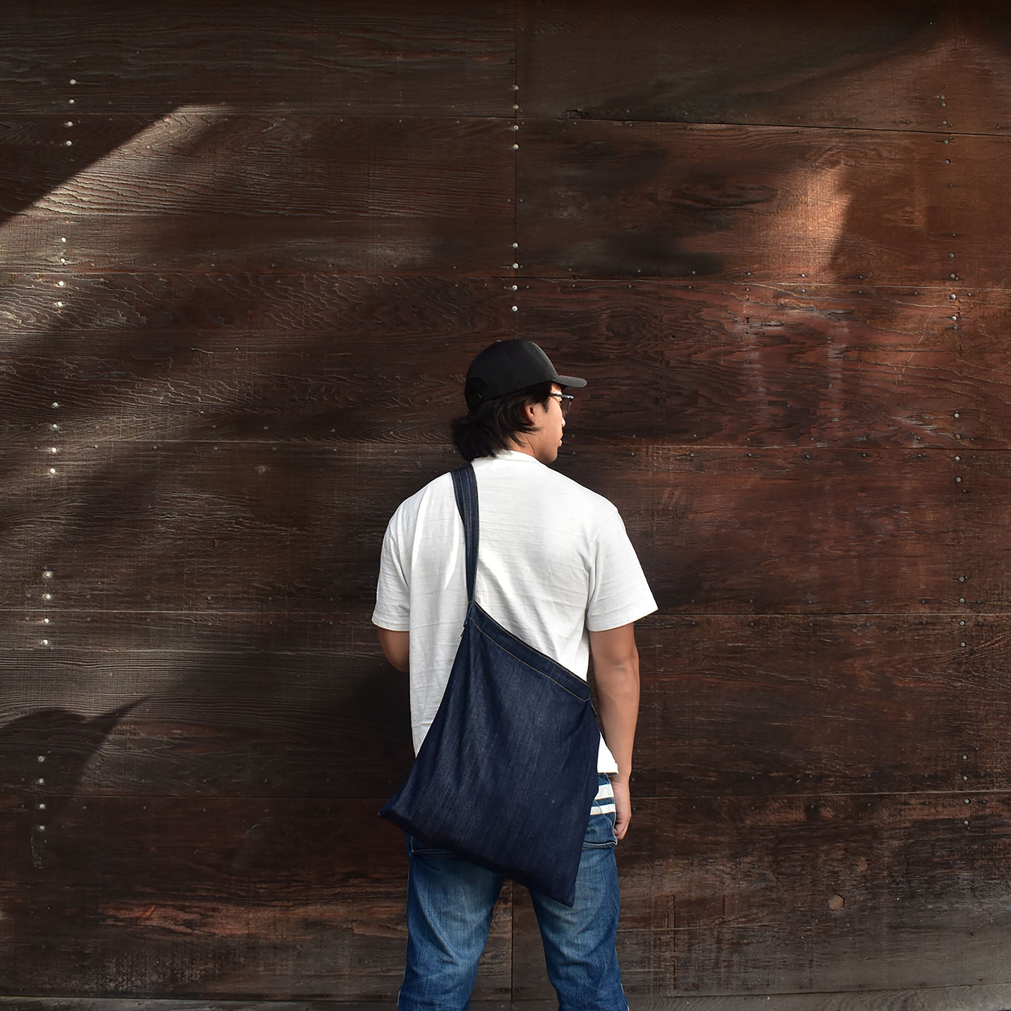 Man wearing a minimalist Japanese denim shoulder bag against a rustic wooden wall, showcasing casual and versatile streetwear style. 