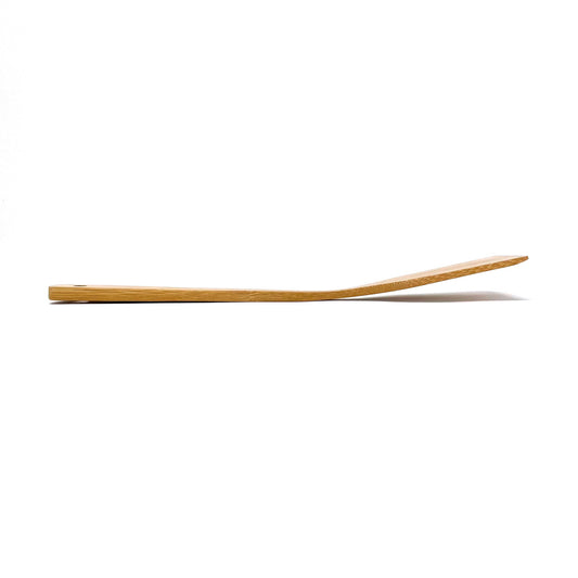 Side profile view of a handcrafted bamboo cooking turner laid flat against a white background, showcasing its slight upward curve designed for optimal leverage and ease of flipping food during cooking.