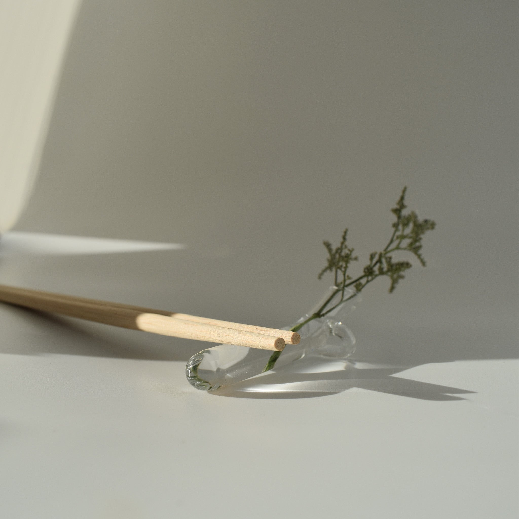 Clear hand-blown glass utensil rest holding wooden chopsticks with a small sprig of greenery, photographed in soft natural light.