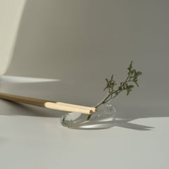 Clear hand-blown glass utensil rest holding wooden chopsticks with a small sprig of greenery, photographed in soft natural light.