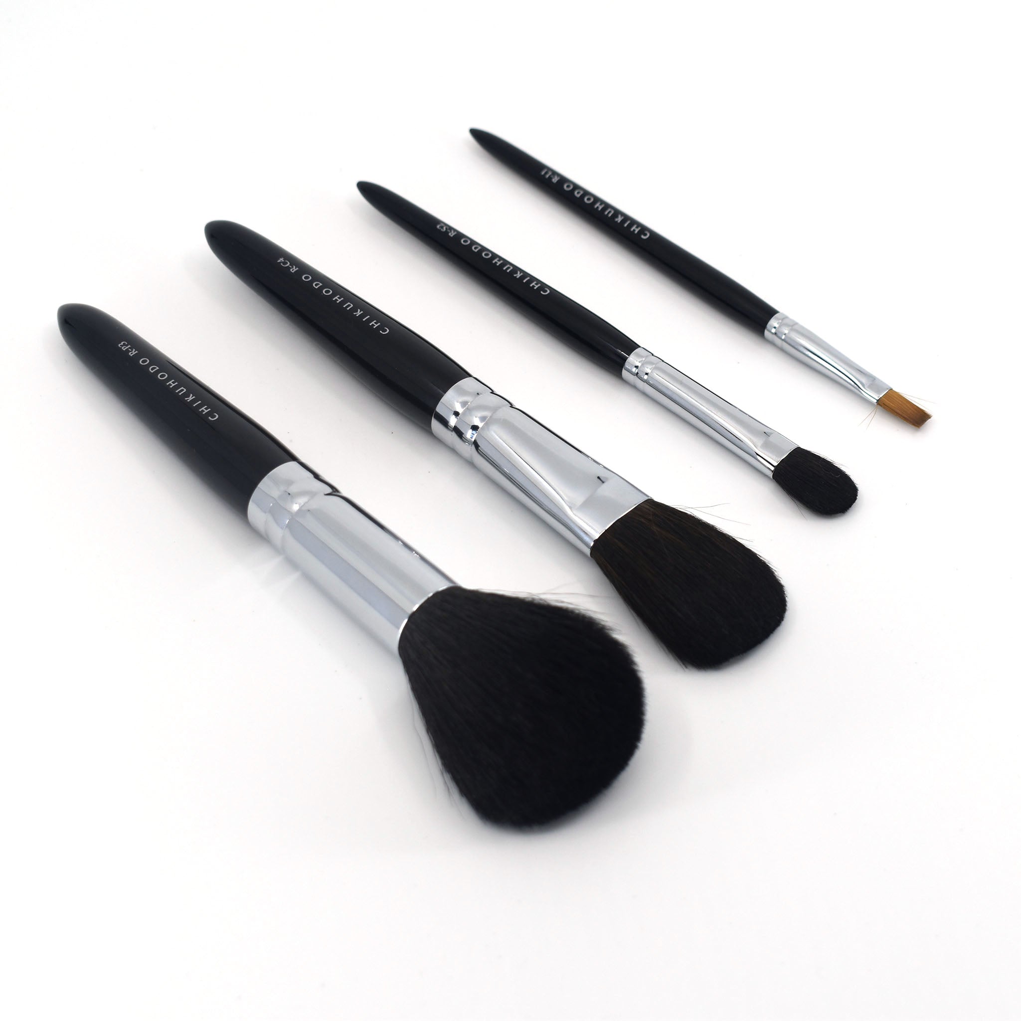 Angled view of four Chikuhodo makeup brushes arranged diagonally on a white surface, featuring sleek black handles, silver ferrules, and soft bristles in varying shapes for face and eye application.