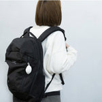 Woman carrying a black backpack with the Cocoon Eco Bag by 100 Percent attached to the front zipper. The compact, oval-shaped white case hangs from the bag, highlighting its convenient clip-on design and stylish minimalism for everyday travel or errands.