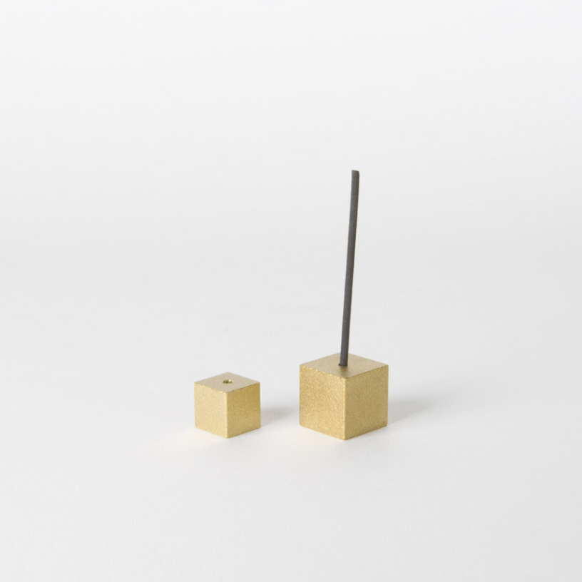 Minimalist Japanese gold brass cube incense holders, one displayed with a burning stick, on a clean white surface.