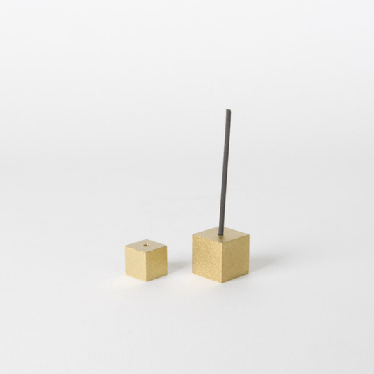 Minimalist Japanese gold brass cube incense holders, one displayed with a burning stick, on a clean white surface.