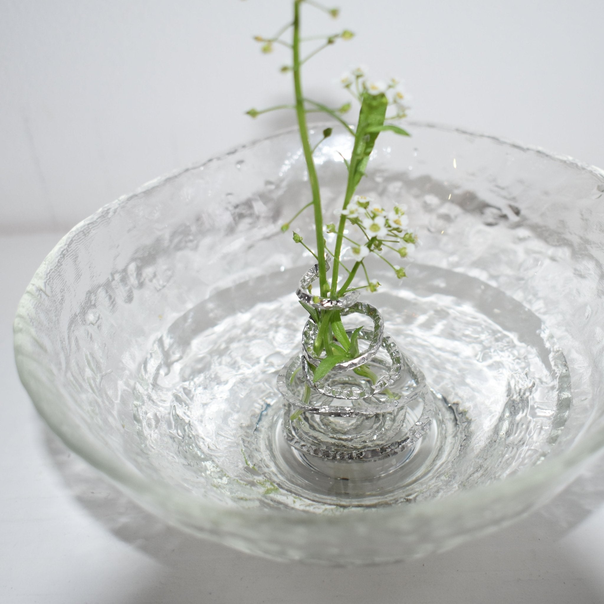 Delicate white flowers held by a sculptural silver spiral flower holder submerged in a rippled glass bowl, showcasing a modern minimalist approach to ikebana and artistic floral design.