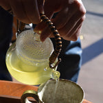 Glass kyusu teapot with rattan handle pouring Japanese green tea into a ceramic cup.