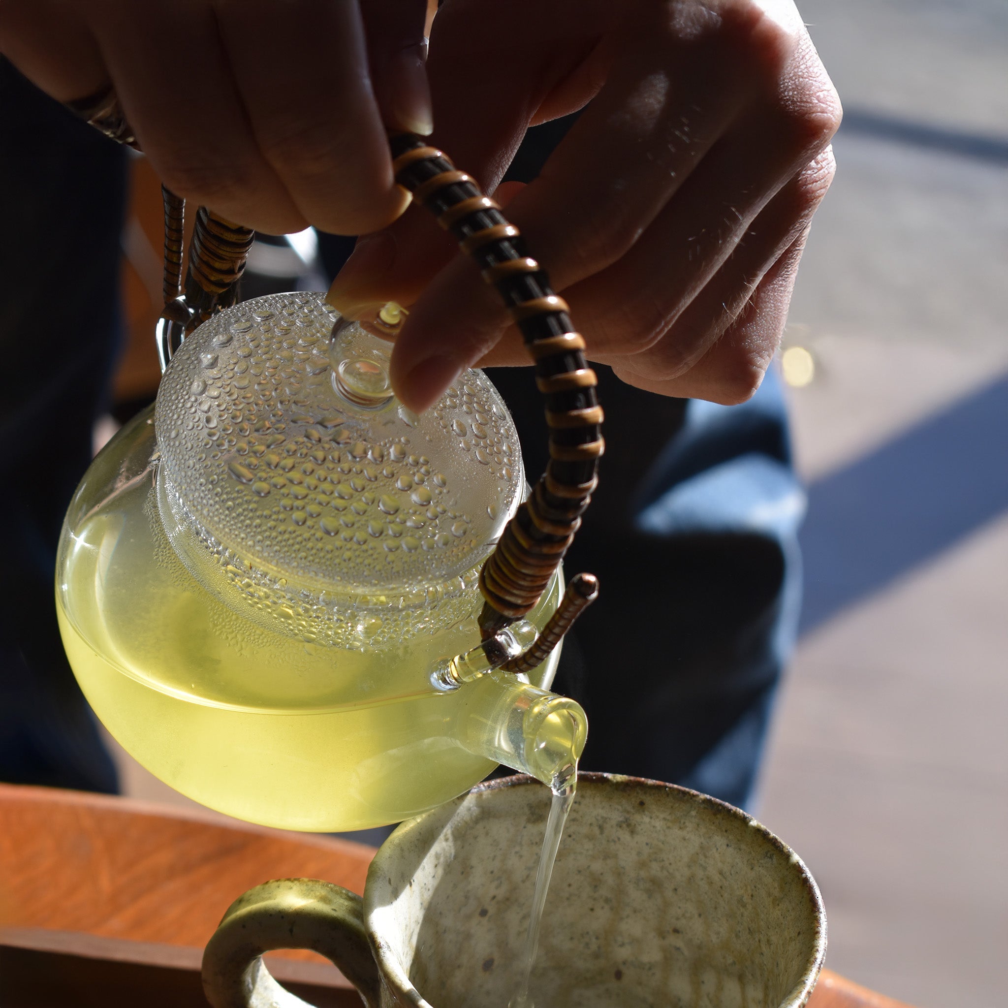 Glass kyusu teapot with rattan handle pouring Japanese green tea into a ceramic cup.