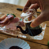 Japanese smooth pour glass soy sauce dispenser held by hand, serving soy sauce alongside sushi and sashimi.