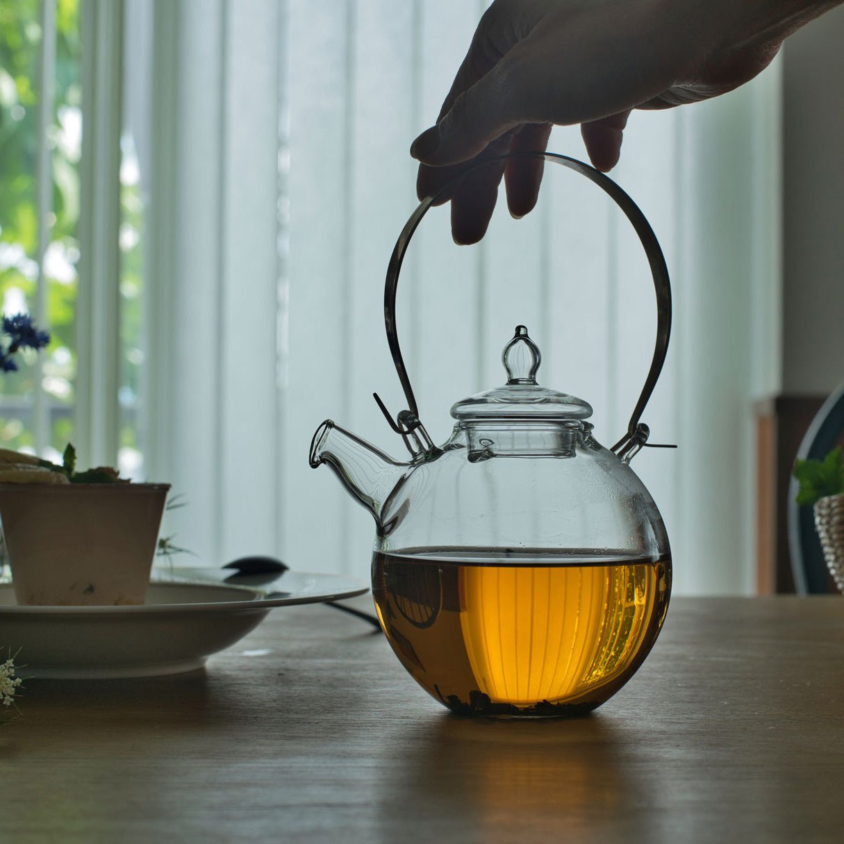 Hand holding the brass handle of a Manmaru glass teapot filled with freshly brewed tea, showcasing its elegant Japanese design on a wooden table.
