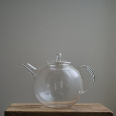 Transparent Manmaru glass teapot with a classic handle and round shape, placed on a wooden surface, ideal for serving tea while showcasing the natural colors of the brew.