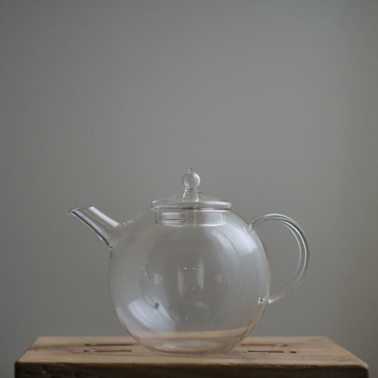 Transparent Manmaru glass teapot with a classic handle and round shape, placed on a wooden surface, ideal for serving tea while showcasing the natural colors of the brew.