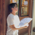 A man cools himself with an open Okayama selvedge denim folding fan by a sunlit window.