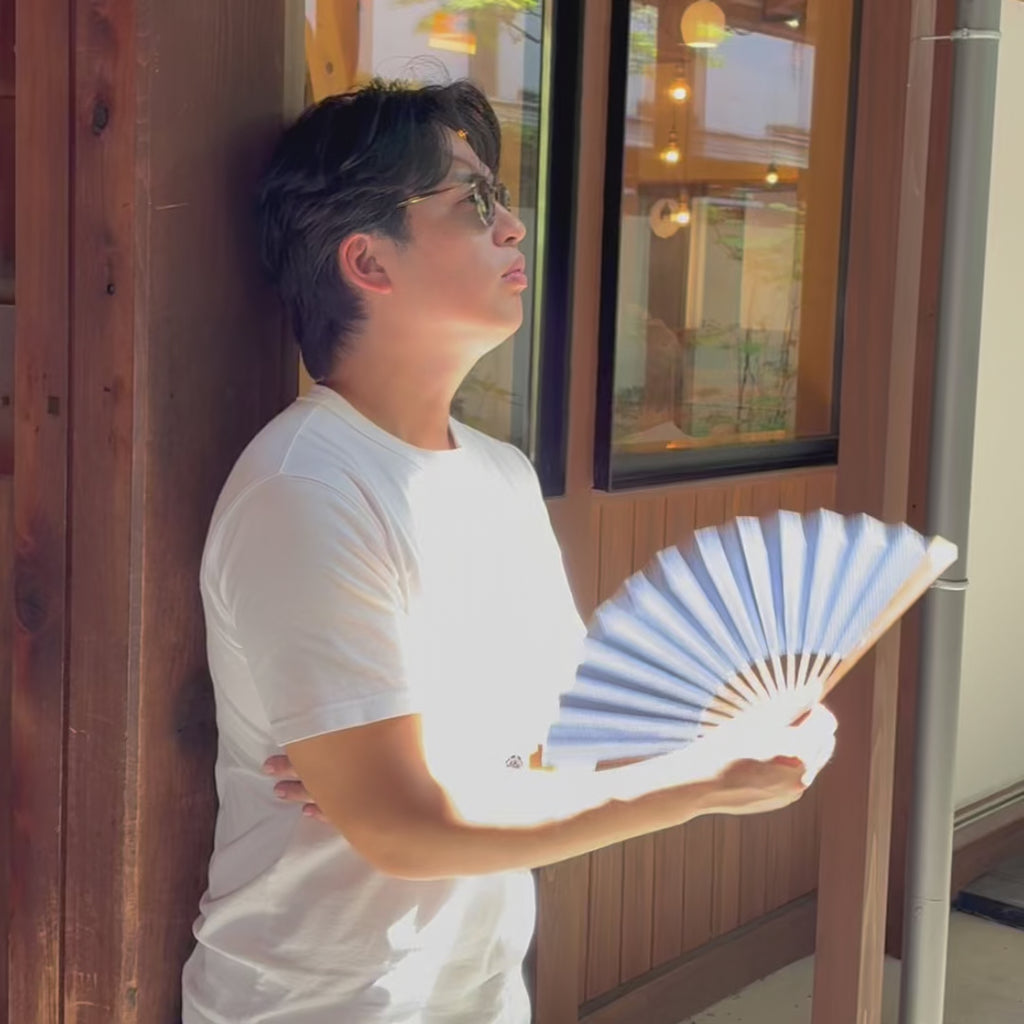 A man cools himself with an open Okayama selvedge denim folding fan by a sunlit window.