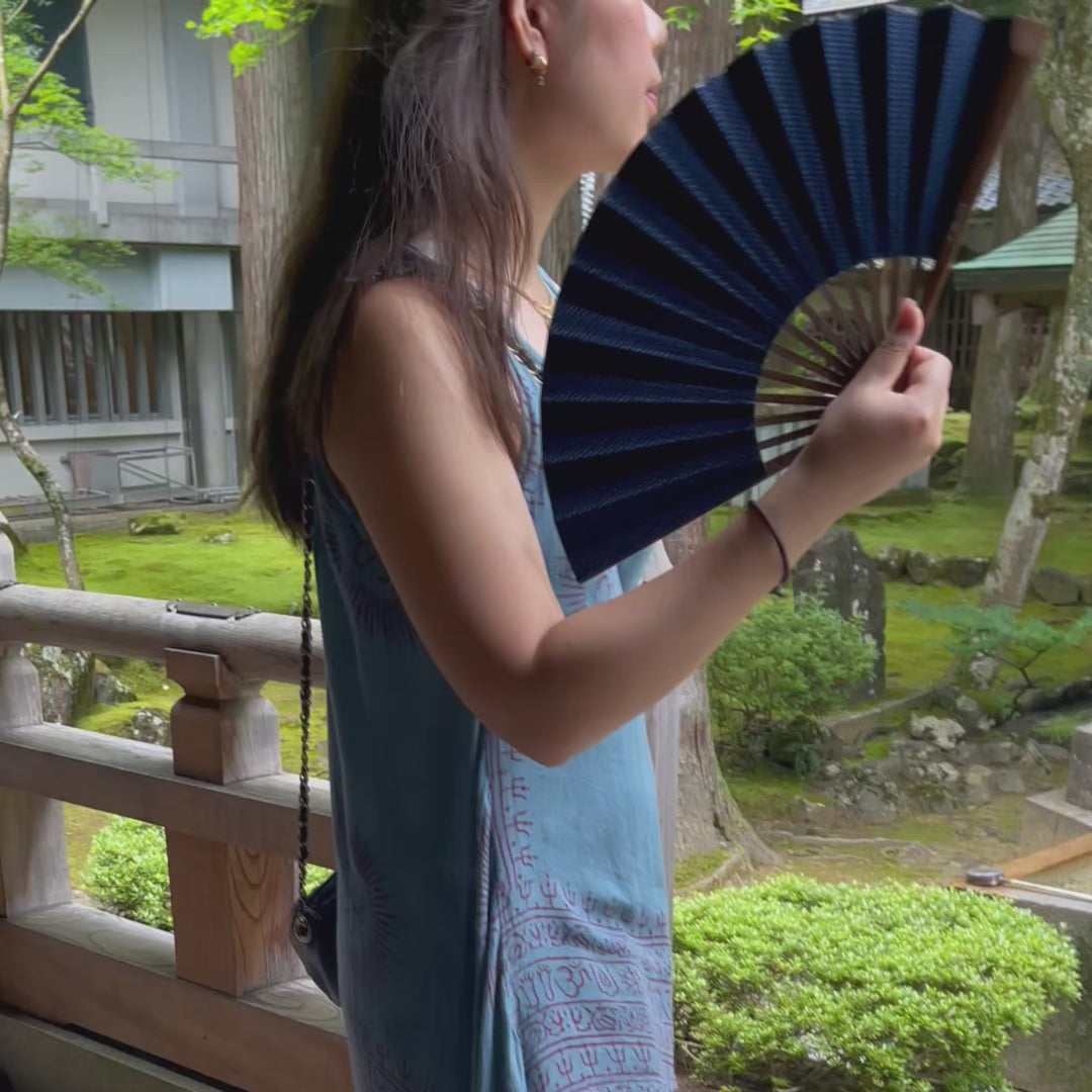 A woman holds an open Sashiko indigo folding fan in a green Japanese garden.